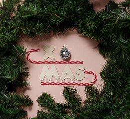 Merry Christmas inscription of wooden letters on a pink pastel background and a caramel cane framed with spruce branches