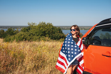 Young woman with the flag of the United States of America standing near near the car on a sunny day in nature