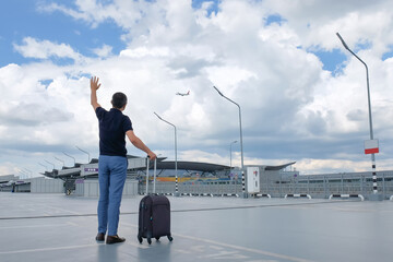 A man passenger with a suitcase in an open airport parking, looks at the sky, sees off the plane. View from the back. Travel concept