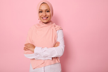 Fototapeta premium Waist-length portrait of a gorgeous Arabic Muslim woman in pink hijab posing looking to camera with attractive gaze, toothy smile, confident look and crossed arms, on pink background with copy space