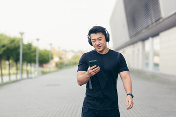 Male asian athlete on a morning run in the park near the stadium listens to music and podcast in big headphones uses the phone