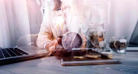 business hand holding lightbulb with using smartphone, laptop computer and money stack in office. idea saving energy and accounting finance concept in morning light