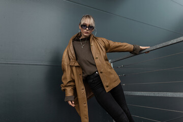 Stylish cool young woman model with fashionable sunglasses in an oversized leather jacket and a sweater with black jeans posing near a metal wall in the city - obrazy, fototapety, plakaty
