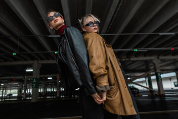 Two fashion models girls in stylish city clothes with a leather jacket with cool sun goggles posing near a parking lot in the city