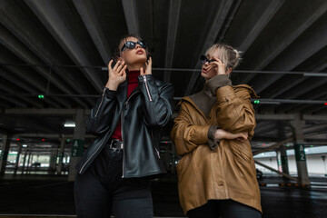 two beautiful young girls in youth style with leather jacket and black jeans with sunglasses posing in a parking lot on the street