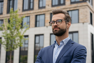 Portrait of confident businessman wearing suit and stylish eyeglasses standing outdoors. Young handsome manager planning project, looking away at copy space. Successful business, career