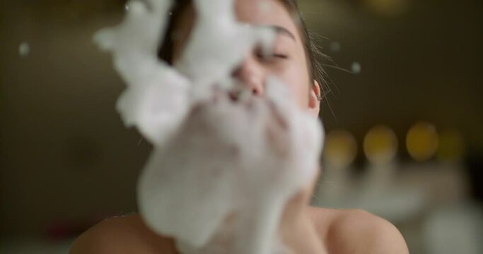 Positive asian girl lying in bathtub blowing soap foam bubbles and laughing. Woman resting in luxury bath doing skincare - wellbeing, happy people 4k footage
