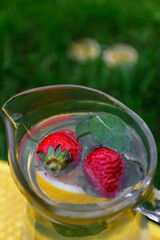 Defocus two strawberry, slice lemon and leaves of mint in glass jug of lemonade on yellow board. Blurred grass background. Pitcher of cool cold drink. Funny smiling face. Out of focus