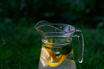 Defocus glass jug of lemonade with slice lemon and mint on natural green background with water drops. Pitcher of fresh cold summer sunny cocktail or punch. Drops. Copy space. Out of focus
