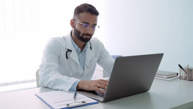 Indian Male Serious Doctor Medical Worker In Modern Clinic Wearing Eyeglasses And White Doctor's Coat Typing Using Laptop Computer Writing Health Personal Data. Telemedicine Healthcare Concept.