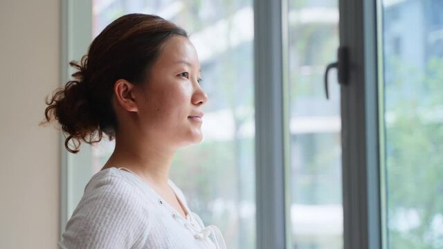 young asian woman stands by the window and looks out the window after getting up at home