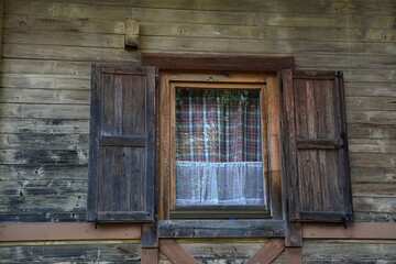 Small window on historical wooden cottage from northern Slovakia with opened window shutters and decorative laced and checkered curtains visible.