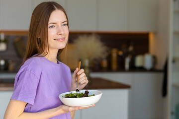 Young and happy caucasian woman eating healthy salad standing in the home kitchen, healthy lifestyle