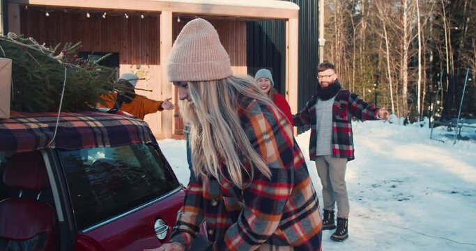 Two young happy women exit red car, hug cheerful multiethnic friends, enter snowy sunny winter chalet house slow motion.
