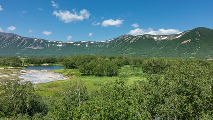 Fototapeta premium The caldera of an extinct volcano is surrounded by a mountain range. In the valley there is a blue lake, a green meadow, thickets of deciduous trees. Clouds in the azure sky. Summer. Kamchatka. Uzon