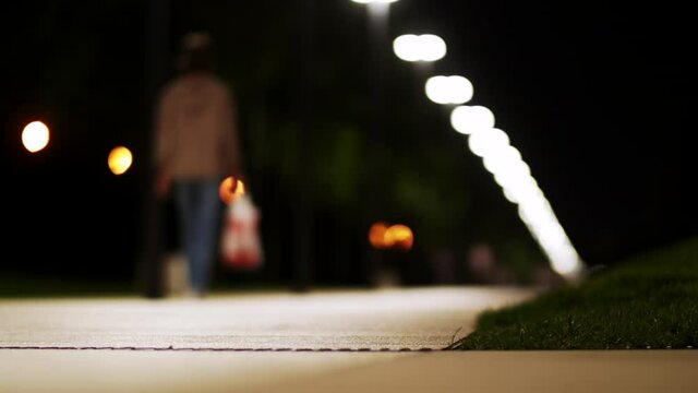 Woman with bag walks on evening alley illuminated by streetlights and man runs asking to give way and people rollerblade
