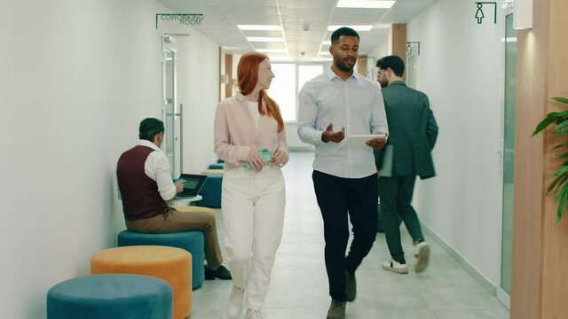 A black man is walking with a beautiful ginger woman in the office, they sit down at a seating area within the office and behind them three people are doing work at coffee tables