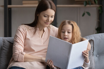 Loving young mother cuddling little kid daughter, reading fairytales or stories in paper book together resting on cozy sofa. Happy two female generations family involved in domestic hobby activity.