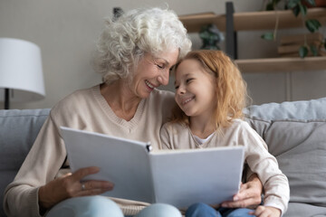 Affectionate happy older senior grandmother reading interesting fairytale stories with joyful small kid daughter in paper book, relaxing together at home, joyful multigenerational family pastime.
