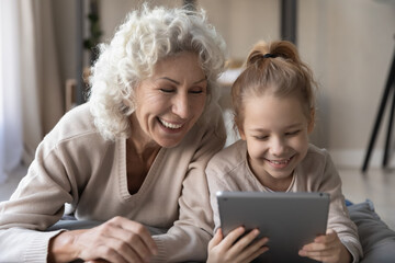 Fototapeta na wymiar Overjoyed caring mature senior grandmother using digital computer tablet with laughing little adorable grandkid girl, lying together on soft pillows, different generations family entertaining online.