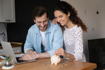 Young married couple in domestic kitchen with laptop put coins in piggy bank, spouses saving money,...