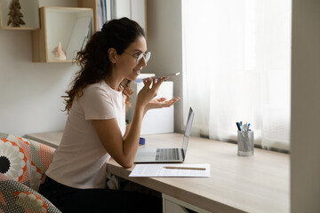Smiling millennial woman in glasses sit at workplace desk in home office holding smart phone...