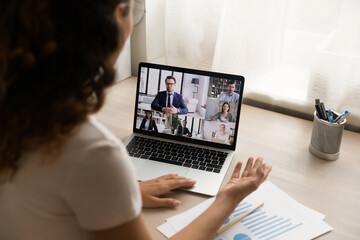 Fototapeta premium Diverse people participate in videoconference make business involved in online remote corporate briefing, pc monitor view over woman shoulder sit at desk attending in videocall. Virtual event concept