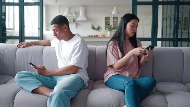 Young upset interracial couple of African American man and Asian woman sitting on the couch back to back, after the quarrel, holding smartphones in hands and turning away from each other.