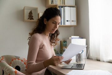 Beautiful Hispanic millennial woman sit at workplace desk reading received correspondence letter,...