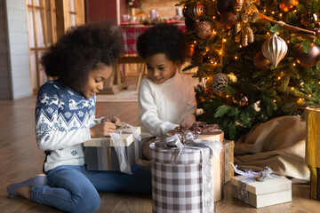 Happy small teen African American children sit on floor near fir tree open gifts presents on Christmas morning. Smiling little biracial kids enjoy winter holidays unpack boxes for New Year.