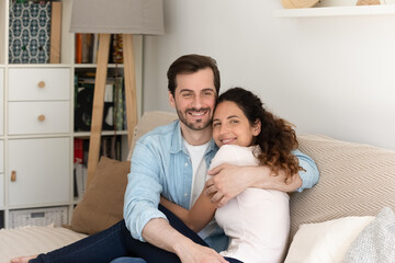 Obraz na płótnie Canvas Couple hugging sit on sofa smile look at camera. Hispanic wife Caucasian husband feeling love embracing enjoy time in modern own or renter home. Romantic dating, home homeowner family portrait concept