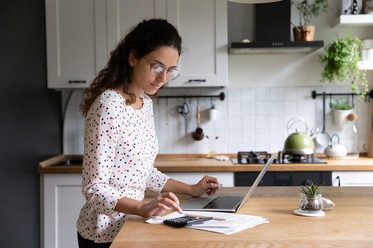 Female standing in home kitchen doing accounting work from home calculates company financial statement, use calculator fill data results on laptop application, housewife manage family budget concept