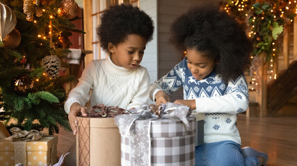 Happy small African American kids feel excited open unbox parcels with gifts presents for New Year. Smiling little teen children unpack box celebrate Christmas together at home. Celebration concept. - obrazy, fototapety, plakaty
