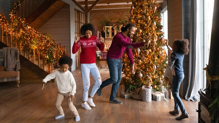 Happy young African American family with kids dance near fir tree in cozy home relax on New Year winter holidays together. Smiling biracial parents with children rest on Christmas play and jump.