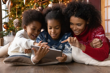 Smiling young African American mother and teen children relax near fir tree on Christmas holiday use pad together. Happy biracial mom and kids speak on video call on tablet on New Year at home.
