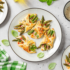 Baked green asparagus with ham and cheese in puff pastry sprinkled with sesame seeds and green basil leaves. Light gray stone background. top view	 - obrazy, fototapety, plakaty