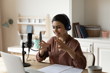 Broadcasting online. Artistic indian woman in headphones sit by desk with laptop speak using...