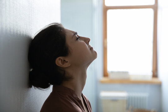 Difficult Life Moment. Tired Stressed Indian Woman Sit Leaning Against White Wall In Light Room Try To Cope With Problems. Side Profile View Of Young Hindu Lady Face With Closed Eyes Think Breath Deep