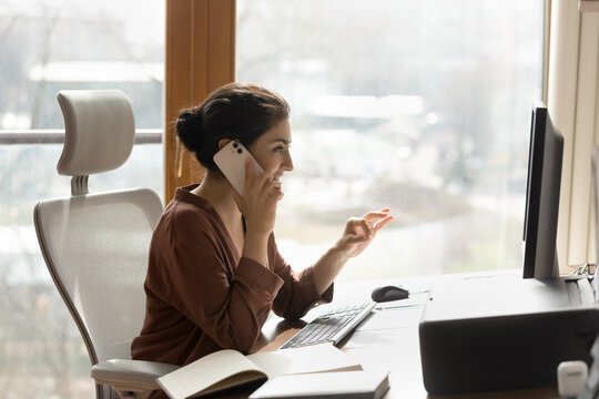 Businesswoman at work. Side shot of confident indian female manager talk on phone at workplace look on computer screen. Smiling young biracial lady expert consult client by cell sale insurance service