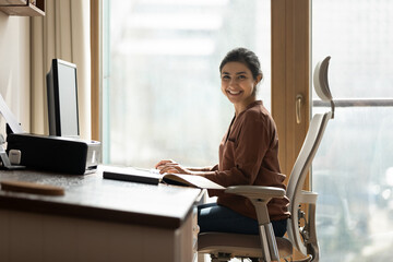 Modern day worker. Portrait of happy biracial business woman freelancer sit by computer at comfy workplace at corporate workspace or at home. Smiling young indian lady office employee look at camera - Powered by Adobe