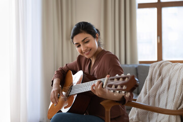 Creative hobby. Talented young mixed race female musician sit in armchair alone compose instrumental song using classic guitar. Smiling biracial lady play calm melody on musical instrument. Copy space - Powered by Adobe