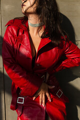 Pretty young woman with wet hair posing in studio, wearing leather red cloak and shiny necklace, natural warm sunset light, standing by wall