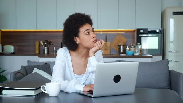 African American ethnicity freelance worker woman working on laptop, typing text, looking up thoughtfully and continues to type text looking at monitor screen. Young woman working, studying remotely