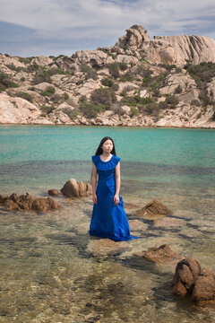 Asian woman in a blue classic dress  standing in the ocean in Sardinia in rocky landscape 