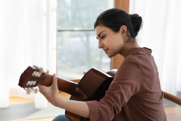 Music of soul. Side view of inspired young indian female guitarist hold acoustic guitar play lyrical melody by window at home. Mixed race lady musician enjoy playing musical instrument arranging song
