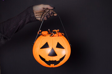 A bucket of candy in the form of a pumpkin lantern Jack on a black background in a hand in a mesh glove. Accessories for Halloween. © Zarina Lukash