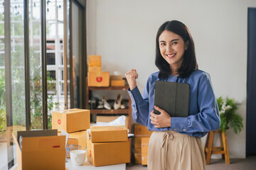 Obraz na płótnie Canvas Success businesswoman happy smiling and holding tablet working at home. Asian woman is owner small business online with shipping box. Concept new normal.
