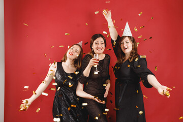 Three delighted women in black dress and party hats over red background with golden confetti. Female party