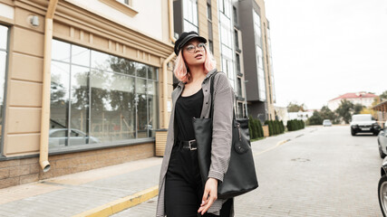 Stylish young beautiful woman with glasses and a hat in fashion clothes with a shirt and black jeans with a bag walks in the city