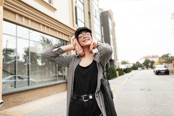 Fashion happy beautiful young woman with a smile in a fashion casual shirt with stylish glasses and a black bag walks in the city